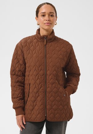 Femme portant une veste matelassée marron avec une fermeture éclair à l’avant, un col montant, des poignets côtelés et des poches latérales, debout devant un fond uni.