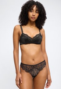 Femme aux cheveux bouclés portant un soutien-gorge en dentelle noire et une culotte assortie en dentelle, debout devant un fond clair uni.