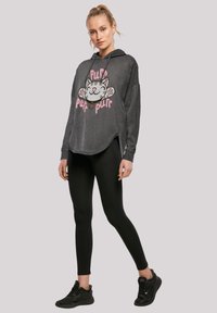 Pelēks kapuces džemperis ar kaķa grafiku un rozā tekstu "purr". Ir brīvs piegriezums, garas piedurknes un noapaļots apakšmalas dizains. Melni legingi un apavi.