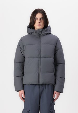 Graue gepolsterte Jacke mit Kapuze, ausgestattet mit einem vorderen Reißverschluss, breiten horizontalen Nähten und einer glatten, matten Textur. Getragen mit lockeren Hosen.