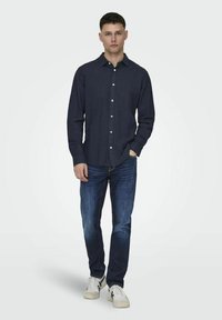 Sötétkék ing, hosszú ujjak, egy zseb, világos gombokkal. Sötét farmernadrággal és fehér sportcipővel viselve. Semleges háttér.