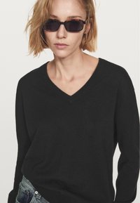 Zwarte v-hals trui van zacht materiaal, met lange mouwen en een relaxte pasvorm. Gemaakt met donkere zonnebril. Achtergrond is simpel wit.