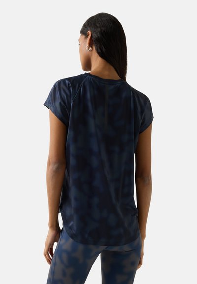 Frau mit langen dunklen Haaren und Creolen, die ein lockeres dunkelblaues kurzärmliges Shirt und passende Leggings trägt, stehend mit dem Rücken zur Kamera.