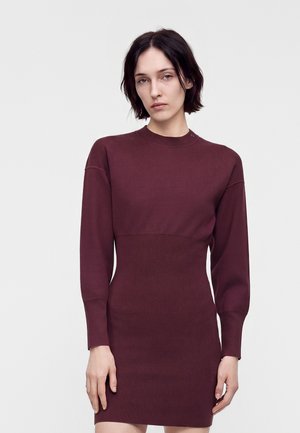 Femme aux cheveux courts et foncés portant une robe en maille ajustée à manches longues de couleur bordeaux foncé, sur un fond clair uni.