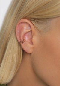 Trije prepleteni uhančki iz rožnatega zlata so nameščeni na zunanji strani ušesa, poudarjajoč gladko, naravno barvo kože in ravne svetle lase.