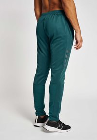 Türkise Sporthosen mit einem elastischen Bund, enganliegenden Bündchen und seitlichen Details. Kombiniert mit schwarzen Sportschuhen mit weißen Sohlen.
