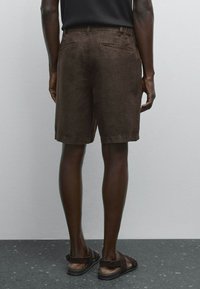 Shorts en lin marron présentant une coupe droite et deux poches arrière, associés à des sandales noires sur une surface grise texturée.