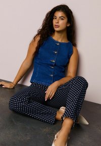 Arany gombokkal díszített kék farmer ujjatlan felső, fekete pöttyös nadrággal párosítva. Világos színű cipő kiemeléssel. Texturált anyag és testhezálló szabás.