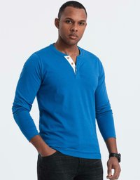 Man som bär en blå långärmad henley-tröja med tre knappar, svarta jeans och en svart klocka på handleden, står med en hand i fickan.