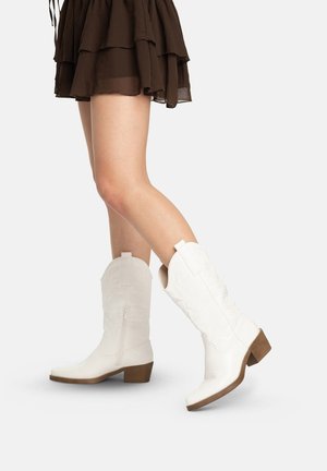 Ben iført lagdelt brun nederdel og hvide cowboystøvler til midt på læggen med lave træhæle mod en ensfarvet baggrund.