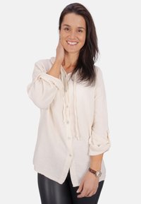 Cremfarbendes, strukturiertes Hemd mit umgeschlagenen Ärmeln und einem Kordelzug-Halsausschnitt, kombiniert mit schwarzen Leggings und einer braunen Uhr.