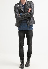 Czarna skórzana kurtka motocyklowa, niebieski sweter z teksturą, czarne dopasowane dżinsy oraz czarne sznurowane buty. Model stoi ze skrzyżowanymi ramionami.