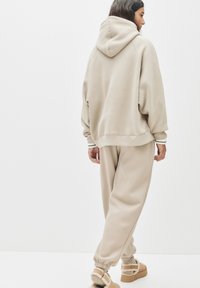 Beige übergroßer Hoodie mit weicher Textur, kombiniert mit lockeren beigen Sweatpants und beigen flauschigen Hausschuhen, von hinten gezeigt.