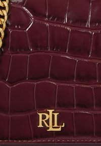 Lähikuva tummasta viininpunaisesta krokotiilinnahkasta, jossa on kultaketju ja kultainen RL-logo-embleemi.