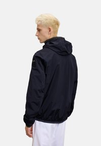 Marineblå vindjakke med hætte, featuring elastiske manchetter og en snoretræk i bunden. Inkluderer et logo patch på venstre skulder.