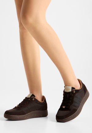 Persona que lleva zapatillas deportivas casuales de color marrón oscuro con suelas moteadas sobre un fondo claro, mostrando el pie izquierdo plano y el derecho levantado.