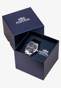 Montre en acier inoxydable argenté avec un cadran bleu marine, fonction date et un bracelet noir, présentée dans une boîte rectangulaire marine avec logo.