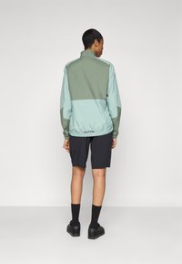 Veste légère bicolore en vert menthe et olive, avec un col montant et un ourlet élastique. Associée à un short noir et des chaussures noires.