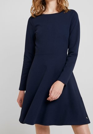Kobieta ubrana w granatową, z długim rękawem, do kolan sukienkę w kształcie litery A na jednolitym szarym tle.