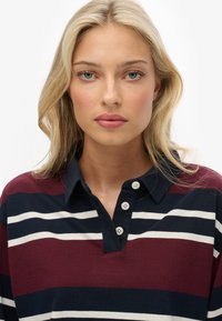 Blond kvinde med blå øjne iført en maroon, navy og hvid stribet rugbytrøje med en navy krave og tre hvide knapper.