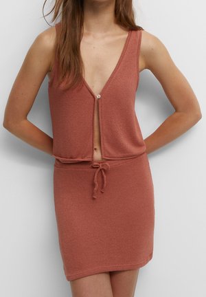 Vrouw draagt een mouwloze, roestkleurige gebreide jurk met een diepe V-hals en een taillekoord, staand tegen een effen witte achtergrond.