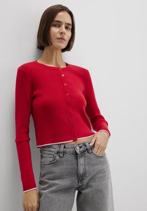 Frau trägt eine eng anliegende rote Strickjacke mit Knöpfen und hoch taillierte ausgebleichte graue Jeans, steht vor einer schlichten weißen Wand.