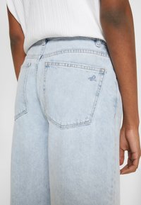 Vaqueros de denim azul claro con un corte relajado, que cuentan con dos bolsillos traseros y detalles de costura sutiles. La tela parece ligeramente desgastada.