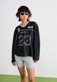 Juodas sportinis ilgomis rankovėmis marškinėliai su baltais akcentais ir numeriu 23, derinami su pilkais Nike šortais. Modelis vilki baltus saulės akinius.