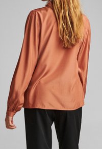 Frau mit langen blonden Haaren, die eine rostfarbene Bluse mit langen Ärmeln und schwarze Hosen trägt, von hinten vor einem schlichten Hintergrund gezeigt.