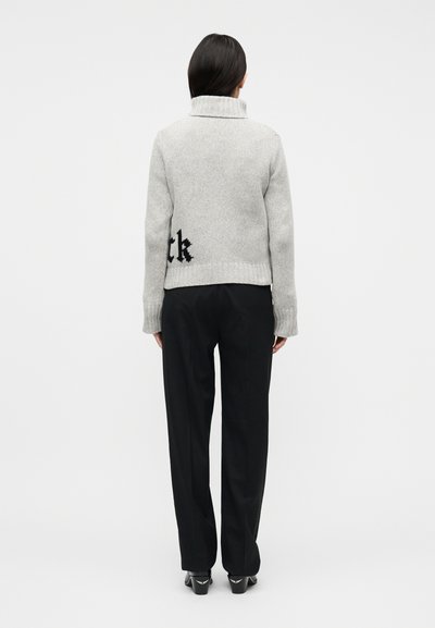 Γυναίκα που φοράει ανοιχτό γκρι πλεκτό πουλόβερ με μερικό μαύρο κείμενο στην πλάτη, μαύρο παντελόνι και μαύρα παπούτσια με τακούνι, στέκεται με την πλάτη προς τα εμπρός.