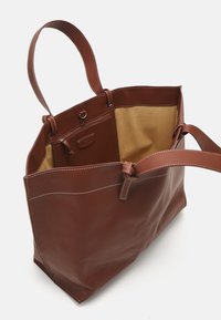 Brown leather tote bag with a smooth texture, two handles, and contrasting beige fabric interior. Features a zip pocket and decorative stitching.