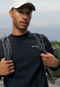 Sweatshirt noir à col rond, arborant un petit logo Columbia blanc. Des sangles en maille grises d'un sac à dos sont visibles sur les épaules.