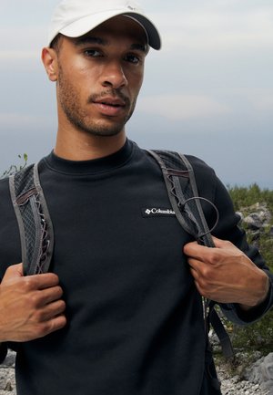 Black sweatshirt with a round neckline, featuring a small white Columbia logo. Gray mesh straps from a backpack are visible on shoulders.