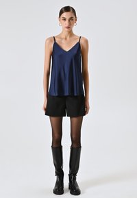 Donna che indossa una canottiera di raso blu navy, pantaloncini neri, collant neri trasparenti, stivali di pelle neri fino al ginocchio e orecchini a cerchio dorati, in piedi su sfondo bianco.