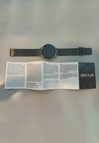 Orologio argentato con quadrante circolare nero, cinturino in rete di acciaio inossidabile e manuale di istruzioni stampato etichettato "SECTOR." Testo e diagrammi visibili.