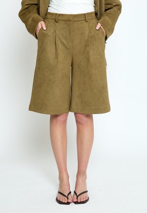 Vrouw draagt olijfgroene, wijde suède shorts, wit topje, bruine jas en zwarte minimalistische sandalen met hak, staat met handen in de zakken.