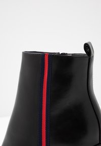Bottine en cuir noir avec un détail en tissu rayé vertical bleu marine et rouge sur le côté et une languette à l'arrière.