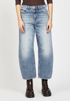 Woman wearing light blue wide-leg cropped jeans and dark brown lace-up leather boots standing against white background.