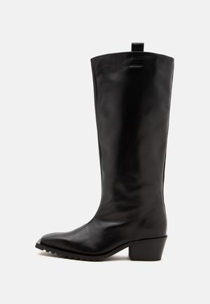 Botte haute en cuir noir avec un talon bas et épais et une languette en haut, vue de côté sur fond blanc.