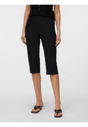 Mujer con pantalones cortos negros a medida hasta la rodilla y top negro sin mangas con sandalias marrones de tacón sobre un fondo liso.