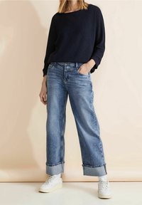 Suéter texturizado azul oscuro combinado con jeans anchos de mezclilla azul claro, con dobladillo acampanado. El modelo lleva zapatillas blancas. El fondo es neutro.