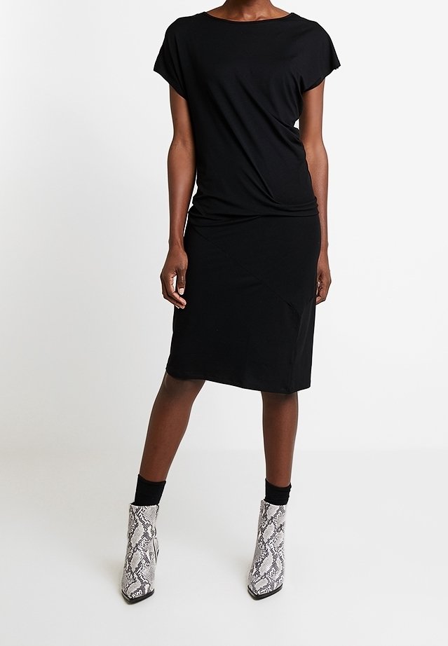Femme portant une robe noire à manches courtes associée à des chaussettes noires et des bottines blanches à motif peau de serpent, debout devant un fond uni.