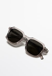 Okulary przeciwsłoneczne z przezroczystą szarą oprawką, prostokątnym kształtem, ciemnymi soczewkami i wyprofilowanymi detalami. Zauszniki mają delikatne wygięcie dopasowujące się do kształtu głowy.