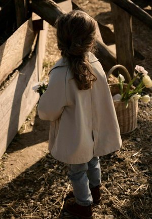 Niño con el cabello trenzado, vestido con abrigo beige y vaqueros, sosteniendo flores blancas cerca de una cerca de madera y una cesta con flores blancas.