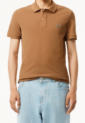 Mann trägt ein braunes Poloshirt mit kurzen Ärmeln und einem bestickten Logo auf der Brust sowie hellblaue Jeans.