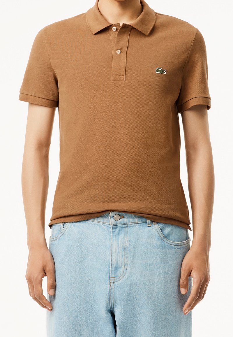Homme portant un polo marron à manches courtes avec un logo brodé sur la poitrine et un jean bleu clair.