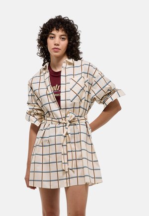 Robe légère de couleur crème avec un motif à carreaux comportant des lignes bleues et brunes, manches retroussées, ceinture nouée à la taille et poche avant.