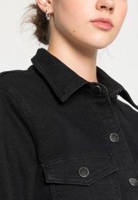 Jaqueta de ganga preta com gola, botões de pressão e bolsos no peito com acessórios em metal prateado. Tecido macio e texturizado.