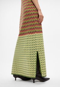 Γυναίκα ντυμένη με ένα μακρύ φόρεμα με ζιγκ-ζαγκ σχέδιο σε πράσινους, μπεζ και κόκκινους τόνους, συνδυασμένο με μαύρες μπότες kitten heel με αιχμηρή μύτη.