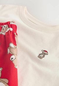T-shirt in cotone color crema con pantaloni corti a fantasia rossa, con grafiche di orsetti teddy. La t-shirt presenta un piccolo orso ricamato con un cappello da Babbo Natale.